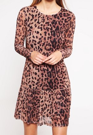 Kobieta ubrana w sukienkę z długim rękawem w panterkę, z okrągłym dekoltem i falbaną na dole, stojąca na tle gładkiej białej ściany.