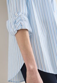 Manche retroussée sur une chemise rayée bleu clair et blanc portée avec un pantalon foncé, montrant le bras et le détail de la patte de boutonnage.