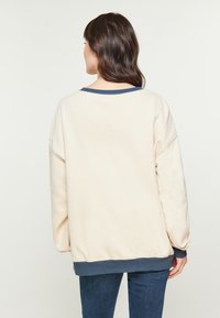 Felpa oversize color crema con colletto e orlo a coste blu navy. Tessuto morbido e cuciture delle spalle abbassate. Il modello indossa jeans blu.