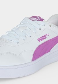 Λευκό αθλητικό παπούτσι Puma με μοβ λογότυπο και ρίγα, λευκά κορδόνια και λευκή σόλα, εμφανιζόμενο από κοντινή πλευρική γωνία.