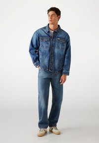 Denim jacket in light blue with two chest pockets, button closure, and a relaxed fit, paired with matching light blue jeans and beige sneakers.