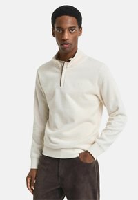 Hellbeige Pullover mit einem Reißverschlusskragen, aus weichem Material gefertigt. Verfügt über lange Ärmel und eine lockere Passform. Kombiniert mit dunkelbraunen Hosen.