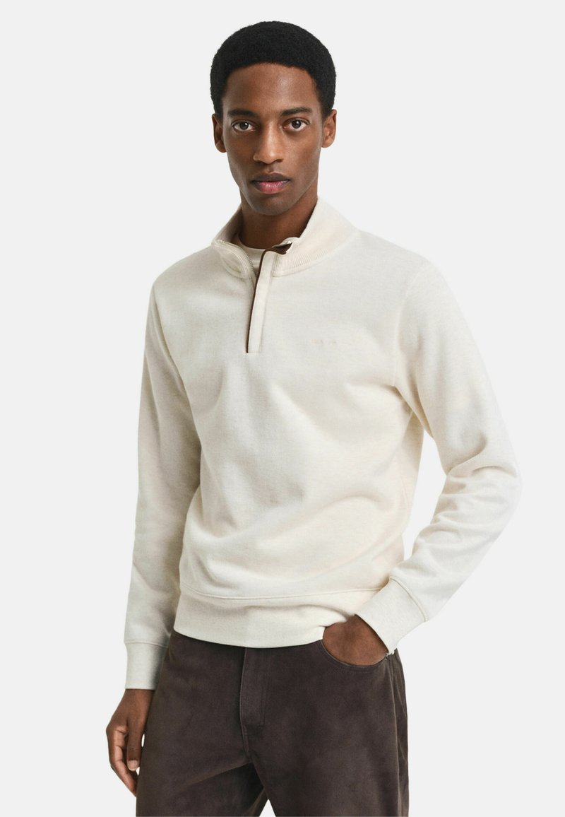 Hellbeige Pullover mit einem Reißverschlusskragen, aus weichem Material gefertigt. Verfügt über lange Ärmel und eine lockere Passform. Kombiniert mit dunkelbraunen Hosen.