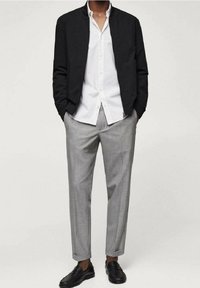 Veste bomber noire portée sur une chemise blanche à boutons, associée à un pantalon gris clair sur mesure et des mocassins noirs. Textures lisses, design moderne.