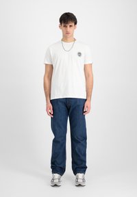 Hvid bomulds-t-shirt med korte ærmer og rund halsudskæring, der har et sort emblem; stylet med mørkeblå denimjeans og grå sneakers.