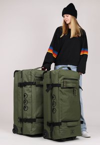 Borse da viaggio verdi in tessuto con accessori neri, ruote a rullo e molteplici tasche con zip; una borsa ha un motivo testurizzato e accenti con il logo.