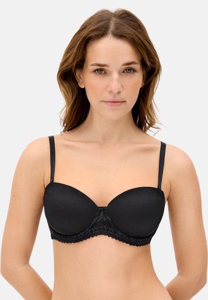 Mujer con cabello castaño hasta los hombros, vestida con un sujetador acolchado negro con tirantes finos y ribete de encaje, de frente contra un fondo liso.