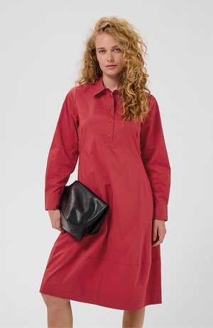 Femme aux cheveux blonds bouclés portant une robe rouge à manches longues tenant une grande pochette noire en cuir pliée sur un fond uni.