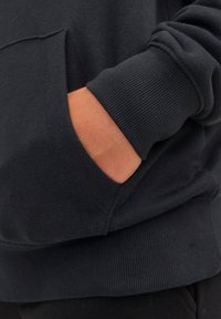 Fekete pulóver kenguruzsebbel, bordázott mandzsettával és strukturált anyagból. A kép közelről mutatja a kezet a zsebben.