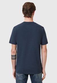 Marinblå bomullst-shirt med rund halsringning och korta ärmar; inga synliga mönster. Bärs med blå jeans och visar en tatuering på handleden.