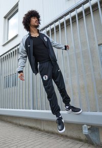 Μαύρο και γκρι αθλητικό κοστούμι Nike με λογότυπο, περιλαμβάνει μια ζακέτα με φερμουάρ και παντελόνια με κορδόνια. Φοριέται με μαύρα αθλητικά παπούτσια Nike. Φόντο σε αστικό περιβάλλον.