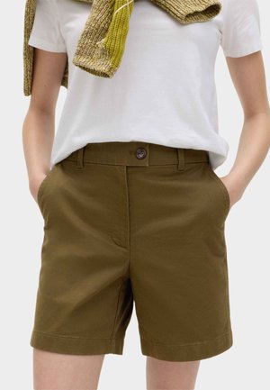 Person iført olivengrønne knapshorts med hænderne i lommerne, hvid t-shirt og en grøn strikket sweater lagt over skuldrene.