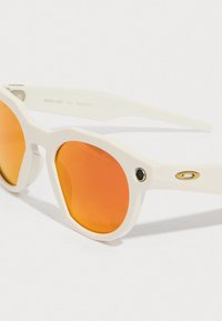 Weiße Sonnenbrille mit orangefarbenen Gläsern, in sechseckiger Form, mit einem goldenen Akzent und einem einzigartigen Design an der Seite des Rahmens.