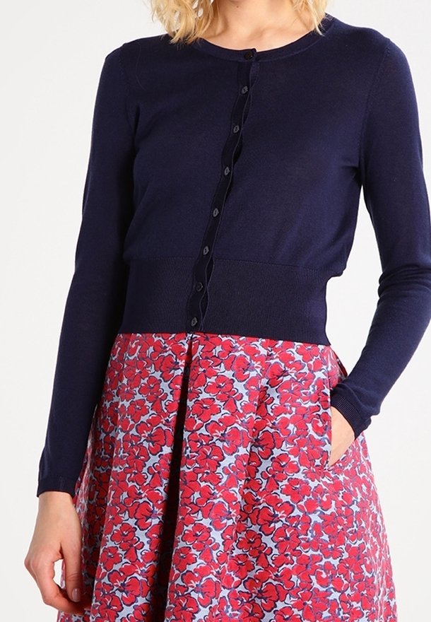 Femme portant un cardigan bleu marine à manches longues et boutonné par-dessus une jupe rouge à fleurs, les mains dans les poches.