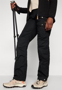 Black outdoor pants with multiple pockets, metal hardware, and seams; paired with beige and orange hiking shoes and trekking poles.