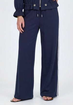 Frau trägt marineblaue, weit geschnittene Hosen mit weißen Seitenstreifen, kombiniert mit Flip-Flops und einer passenden marineblauen Jacke mit goldfarbenem Reißverschluss und Knöpfen.