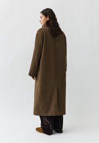 Langer, übergroßer brauner karierter Mantel mit geradem Schnitt, aus leichtem Material und ohne sichtbare Verschlüsse. Getragen über dunklen Hosen und Slippern.