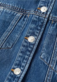 Tissu en denim bleu foncé avec une texture légèrement délavée. Comporte des boutons-pression argentés et des coutures contrastées orange. Vue en gros plan.