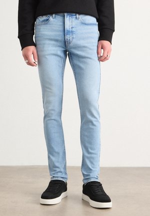 Personne portant un jean slim bleu clair, un haut noir à manches longues, des baskets noires avec semelles blanches et des bagues argentées aux deux mains, debout sur un sol gris.
