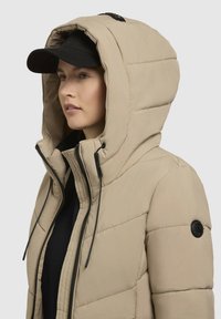 Beige oppustelig jakke med stor hætte, sort lynlås, snore og en tekstureret overflade. Har et rundt sort logo på ærmet.