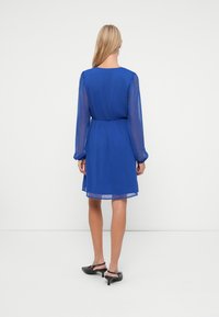 Femeie stând cu spatele, îmbrăcată într-o rochie lungă albastră cu mâneci transparente și pantofi slingback negri cu toc mic, pe un fundal alb.