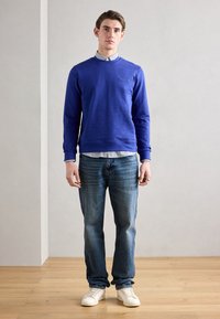 Felpa blu con collo rotondo, tessuto texturizzato e logo frontale. Indossata sopra una maglietta a righe, abbinata a jeans blu e sneakers bianche.