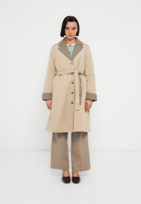 Trench beige avec un col texturé, de gros boutons et une ceinture à nouer. Les poignets à motif à carreaux marron mettent en valeur les manches.