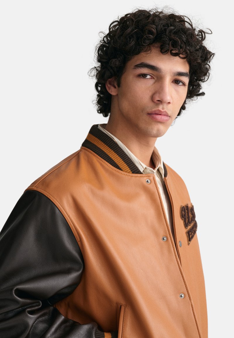 Braune Leder-Collegejacke mit dunklen Lederärmeln, geripptem Kragen, Druckknopfverschluss und dekorativer Stickerei auf der Vorderseite.