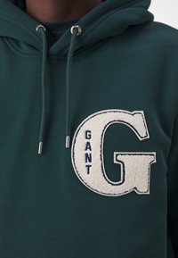 Sweat à capuche vert foncé avec cordons et un grand patch blanc texturé en forme de lettre "G" avec "GANT" brodé verticalement à l'intérieur.