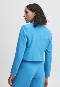 Zkrácený modrý blazer vyrobený z hladké látky s dlouhými rukávy a strukturovaným límcem. Obsahuje upravené švy a minimalistický design.