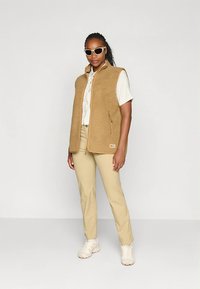 Bež flis brezrokavnik z visokim ovratnikom in žepom z zadrgo, nošen čez svetlo srajco in kombiniran z bež hlačami ter belimi supergami.