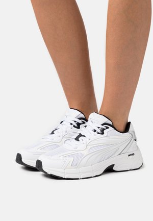 White and black athletic sneakers worn on feet, featuring mesh and synthetic overlays with thick cushioned soles.