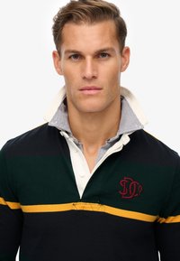 Zwart, groen en geel gestreepte rugbyshirt met een witte kraag. Heeft een geborduurd logo op de borst en een gestructureerde gebreide oppervlakte.