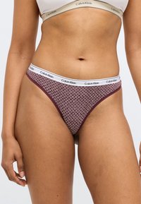 Μπορντό στρινγκ Calvin Klein με λευκή λογότυπη ζώνη και μικρό γεωμετρικό σχέδιο καρδιάς. Κατασκευασμένο από μαλακό ύφασμα με λεία υφή.