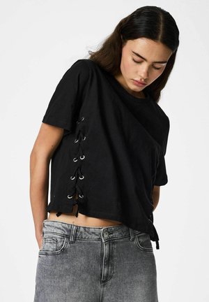 Młoda kobieta w luźnej czarnej koszulce z wiązanymi bokami i wyblakłych szarych dżinsach, patrząca w dół na tle jednolitej białej ściany.