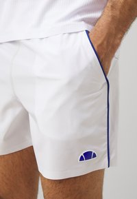 White athletic shorts with a smooth texture, featuring blue accent piping and a small blue and white logo on the left side.