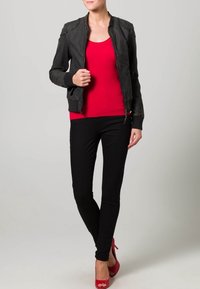 Zwarte leren bomberjack met ribbelmanchetten, rode aansluitende top en zwarte skinny jeans. Model draagt rode hoge hakken. Effen achtergrond.