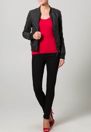 Czarna skórzana kurtka bombowiec z żebrowanymi mankietami, dopasowana czerwona bluzka i czarne skinny jeans. Modelka ma na sobie czerwone szpilki. Gładkie tło.