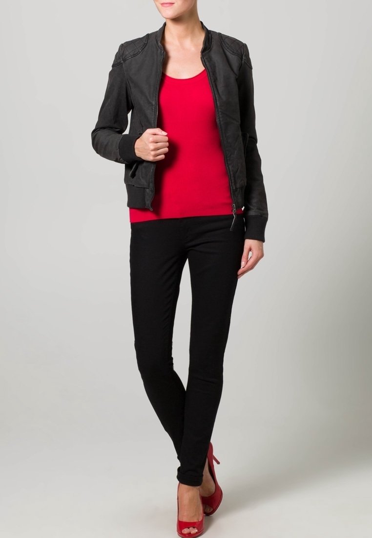 Zwarte leren bomberjack met ribbelmanchetten, rode aansluitende top en zwarte skinny jeans. Model draagt rode hoge hakken. Effen achtergrond.