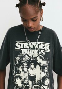 Crni oversized majica s grafikom "Stranger Things" u bijeloj boji, koja prikazuje likove na tamnoj, dramatičnoj pozadini. Srebrni dodaci u obliku nakita.