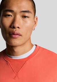 Koraalkleurige sweatshirt met ribbelboord en accentstiksels, gelaagd over een wit shirt. Soepele textuur; close-up van het gezicht van een mannelijke model.