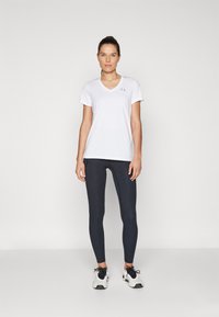 Camicetta bianca a maniche corte con scollo a V e leggins scuri con una texture liscia, abbinati a scarpe sportive grigie con lacci neri.