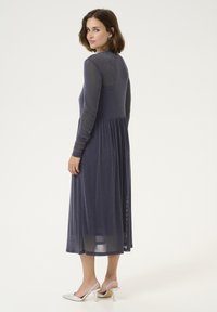 Mörkblå klänning med lång ärm i glittrande tyg, figurnära överdel och flödande kjol. Tillsammans med vita klackskor, minimalistiskt hår. 