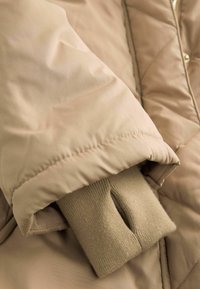 Beige quiltet jakke med blødt, glat stof, ribbestrikkede manchetter og et enkelt design med synlige sømdetaljer.