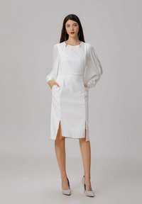 Robe blanche avec des manches bouffantes, des coutures verticales, des fentes latérales et des poches. Portée avec des talons hauts de couleur claire sur un fond neutre.