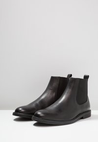 Bottines en cuir noir avec des panneaux élastiques sur les côtés, des bouts ronds et des semelles plates. Fini lisse avec des détails de design minimalistes.