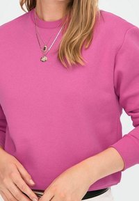 Sudadera rosa hecha de tela suave, con escote redondo y mangas largas. Como accesorios, lleva collares plateados en capas.