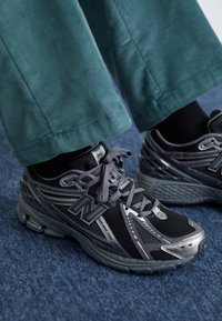 Chaussures de sport noires et grises avec une tige en maille, un bout renforcé et une semelle en caoutchouc texturée, associées à un pantalon en velours côtelé bleu sarcelle.