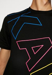 Muž v černém tričku s velkými geometrickými obrysy v modré, růžové a žluté barvě na hrudi a rameni.