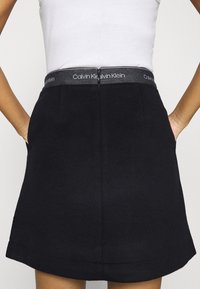 Gonna nera con un design strutturato, caratterizzata da una vita grigia con il logo Calvin Klein. Il tessuto sembra liscio e leggero.